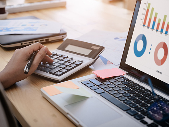 hand using calculator next to laptop displaying budget graphs